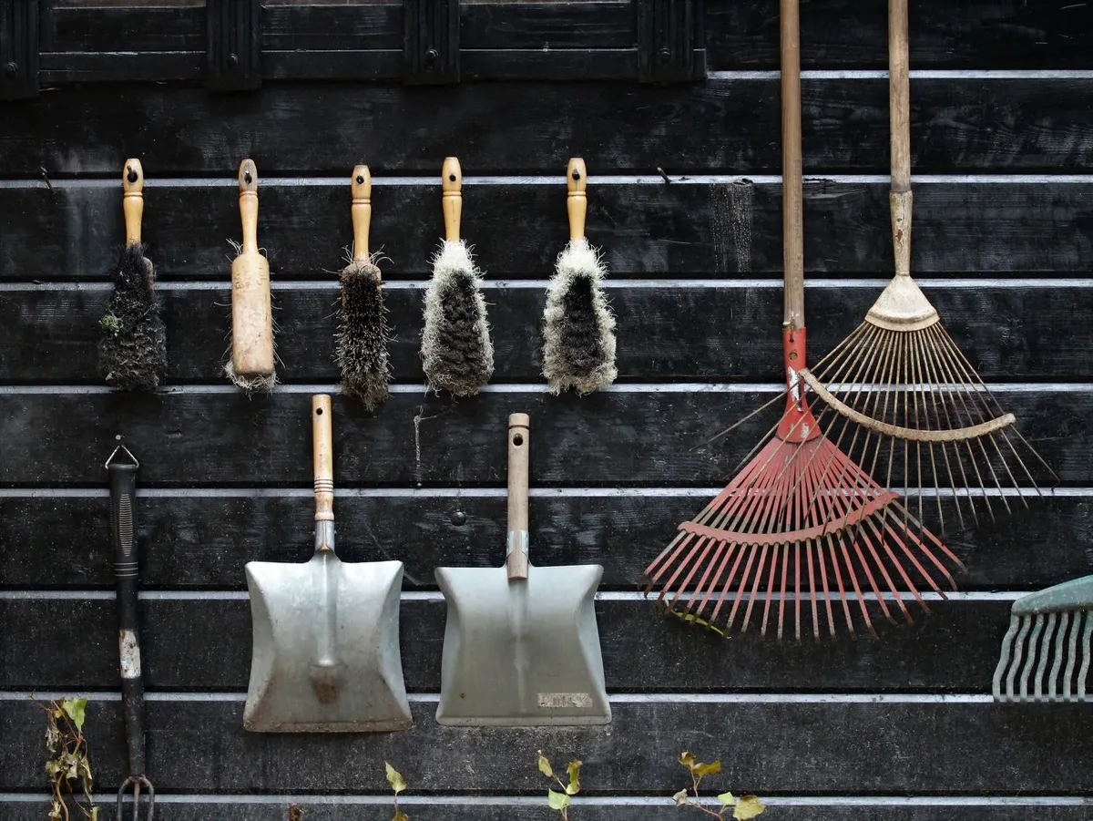 Outils de jardin suspendus sur le bardage en bois sombre d'un abri : balayettes, pelles, râteaux