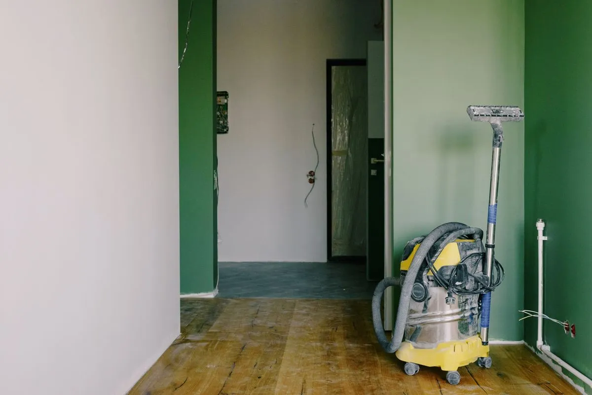Pièce en travaux au parquet ancien usé, avec un aspirateur de chantier jaune posé contre un mur vert