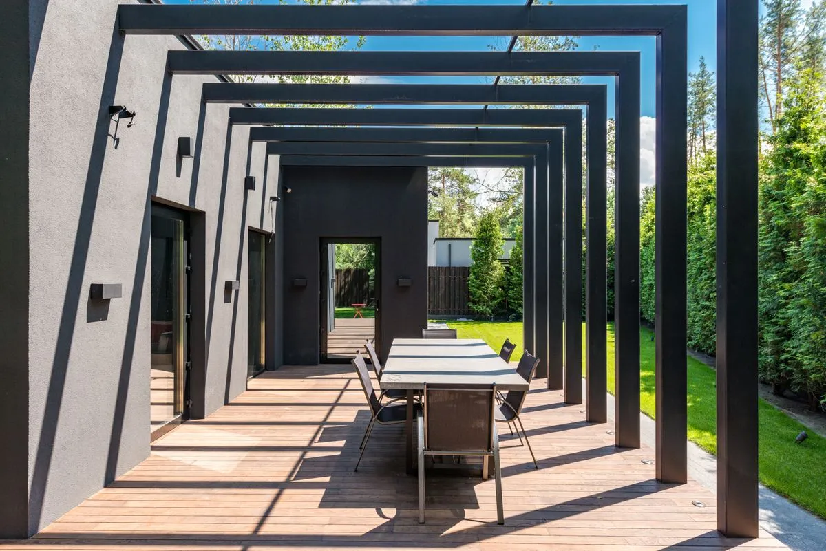 Pergola contemporaine à poteaux et poutres noirs au-dessus d'une terrasse en bois avec table, le long d'une pelouse