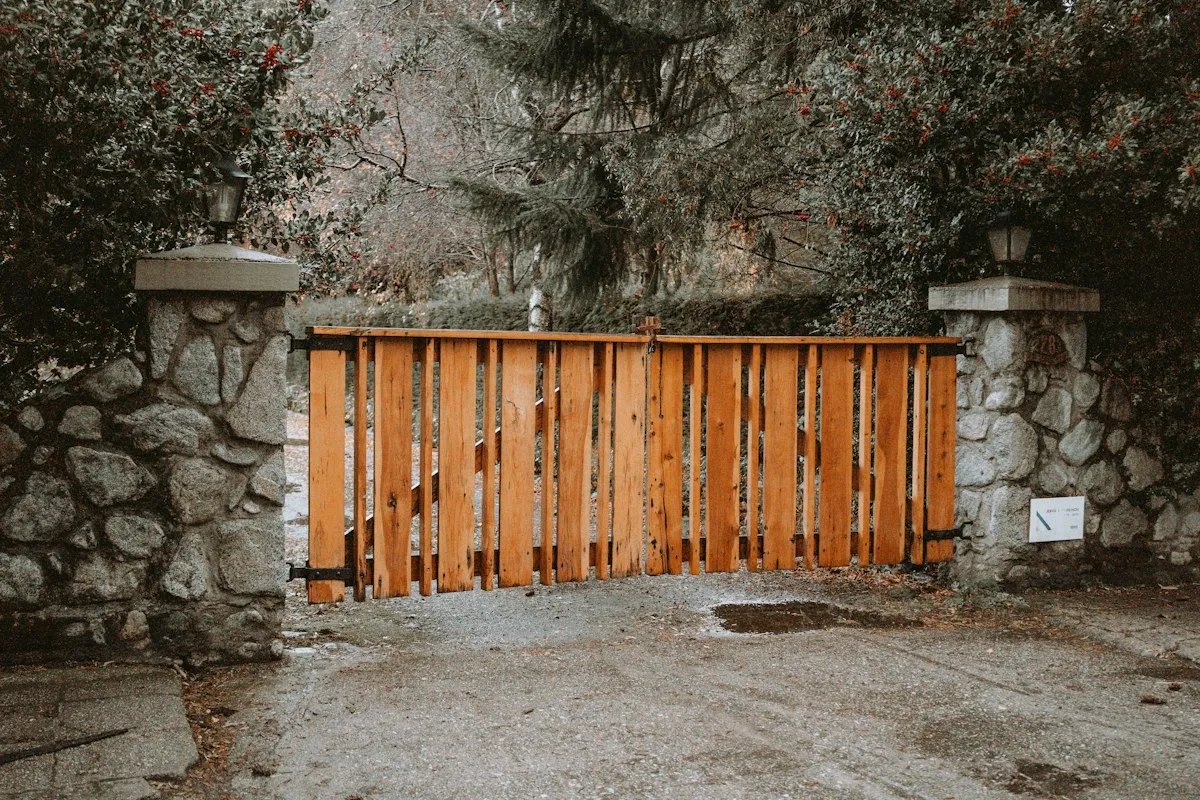 Portail en lames de bois naturel fermant une allée gravillonnée, entre deux piliers en pierre surmontés de lanternes