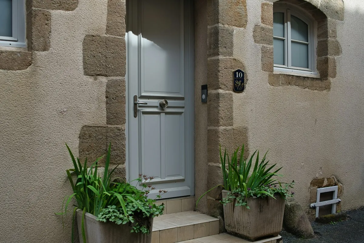 Porte d'entrée pleine peinte en gris clair dans une façade enduite à chaînes d'angle en pierre, avec deux plantes en pot sur le seuil