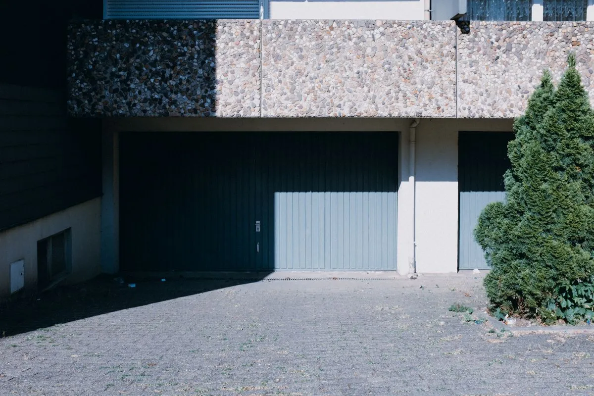 Rangée de portes de garage basculantes en métal rainuré au pied d'un immeuble des années 1970, devant une cour pavée