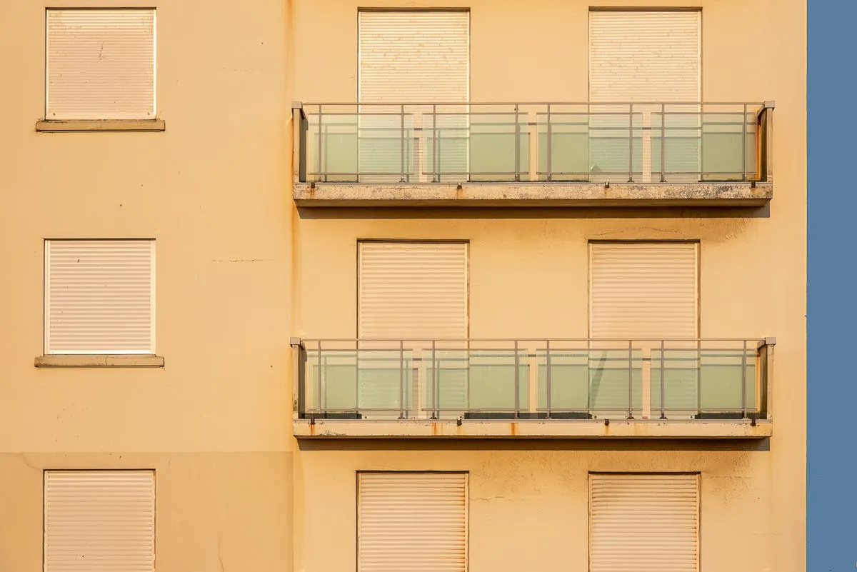 Façade ocre d'immeuble en plein soleil, volets roulants beiges fermés à toutes les fenêtres et balcons vitrés